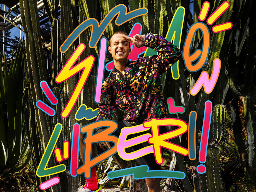 A professional horizontal crop of the adidas Originals Pride Collection campaign featuring Sangiovanni. The shot is cut at the waist, focusing on the artist's torso and expression as he poses with a military salute among cacti. Vibrant, Keith Haring-inspired pop-art graphics by Mauro Bosoni, including the multicolored text "SIAMO LIBERI!!", wrap around his body. Sangiovanni wears the colorful Pride bomber sweatshirt. The background of giant green cacti is illuminated by bright, high-contrast natural light. Photo by Alvise Guadagnino.