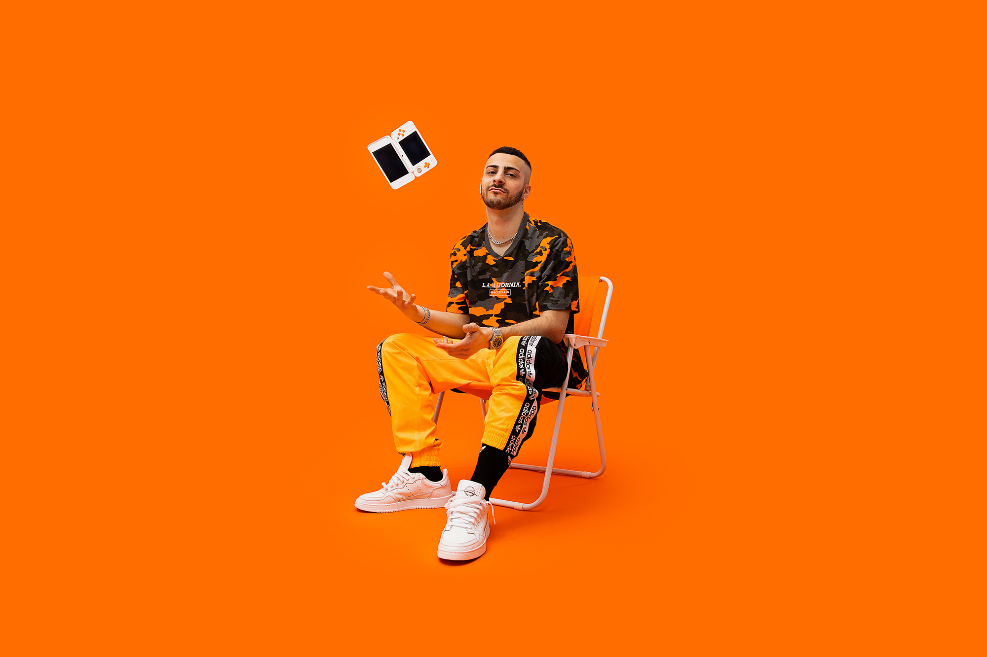 adidas ozweego charlie charles orange background gaming by alvise guadagnino_03 Music producer Charlie Charles sits on a white folding chair against a vibrant orange background for the Adidas Classic Ozweego campaign. He is wearing a camouflage T-shirt, bright orange Adidas track pants and white Ozweego sneakers, with a handheld gaming console floating in the air. Professional fashion photography by Alvise Guadagnino for Adidas Originals.